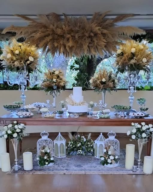 Nuvem de capim dos pampas sobre a mesa do bolo, entre castiçais de cristal, gaiolas brancas e velas — arranjo em tons de areia e branco.